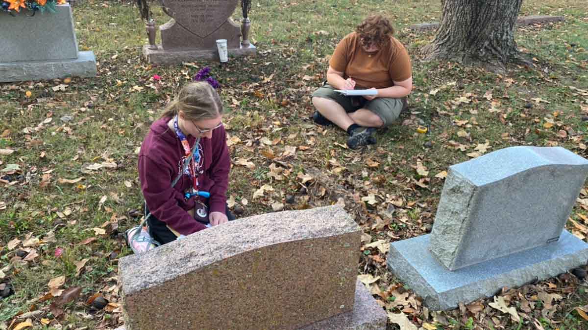 Two students sitting in cemetery measuring head stones