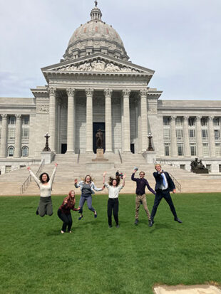College students jumping in the air in front of a state capitol building.