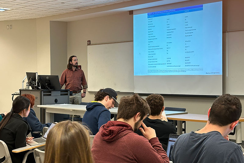 Man standing at classroom podium reviewing a lecture slide