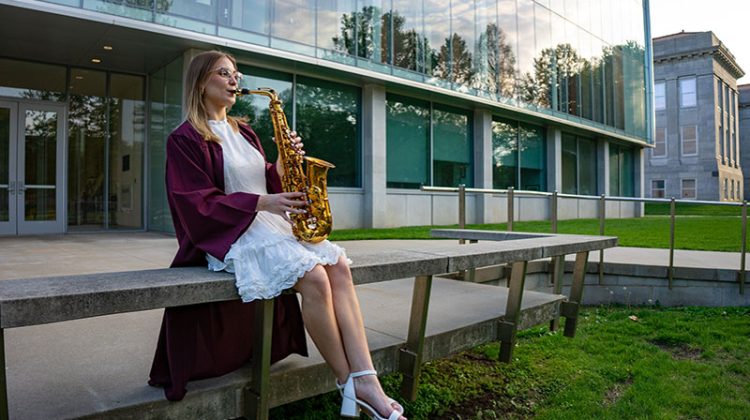 Young woman sitting in front of building in graduation regalia and playing a saxophone
