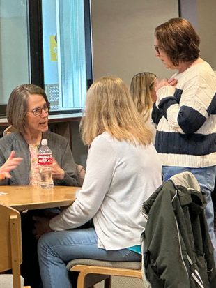 A woman stands at a table and listens to two other women as they speak.