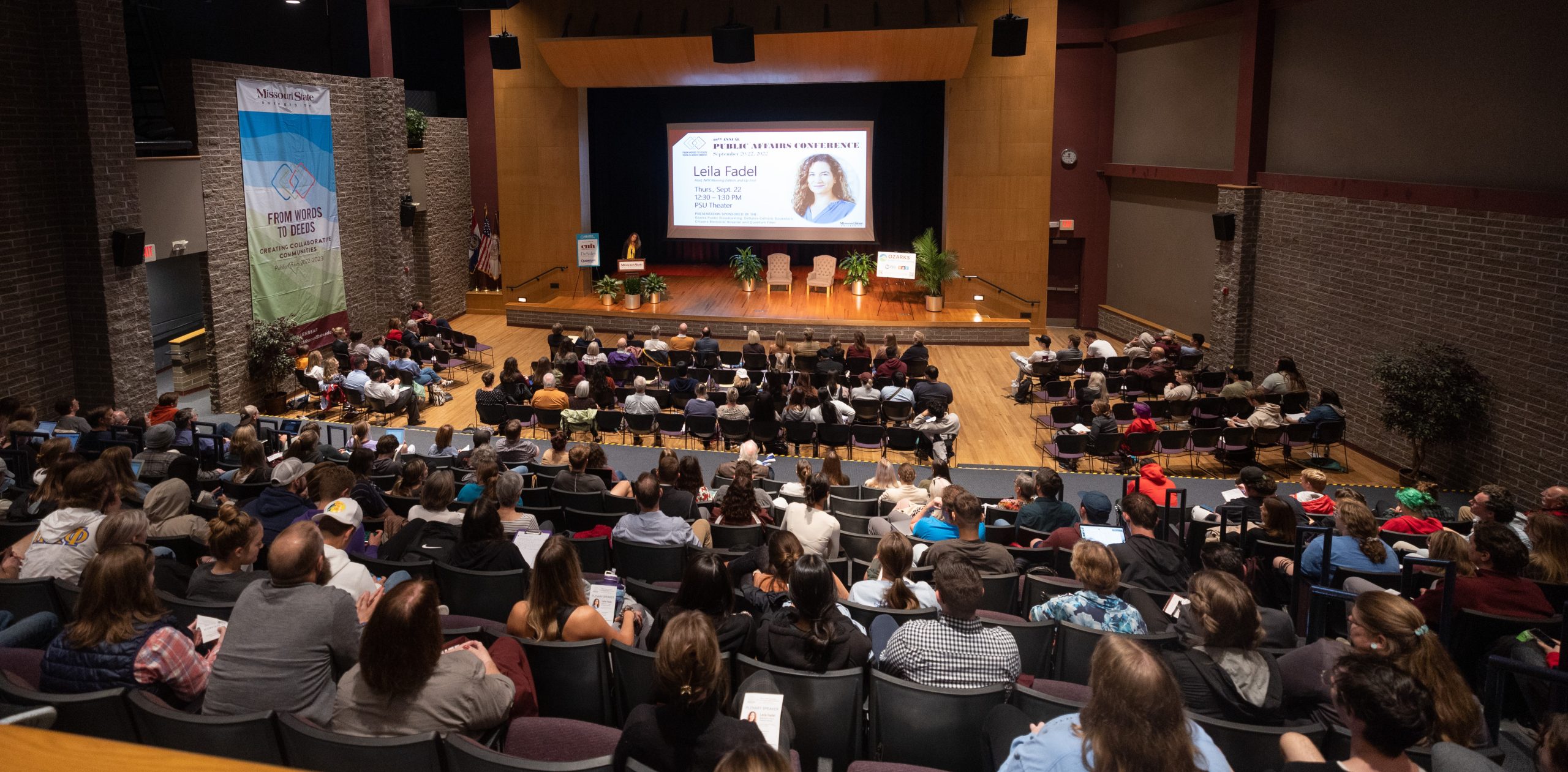 A crowded theater listens to a speaker on stage